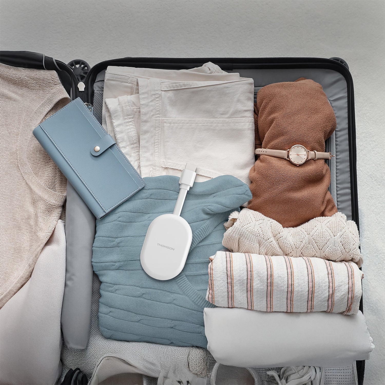 Open suitcase with folded clothes, a streaming dongle, and a blue pouch on a light gray background